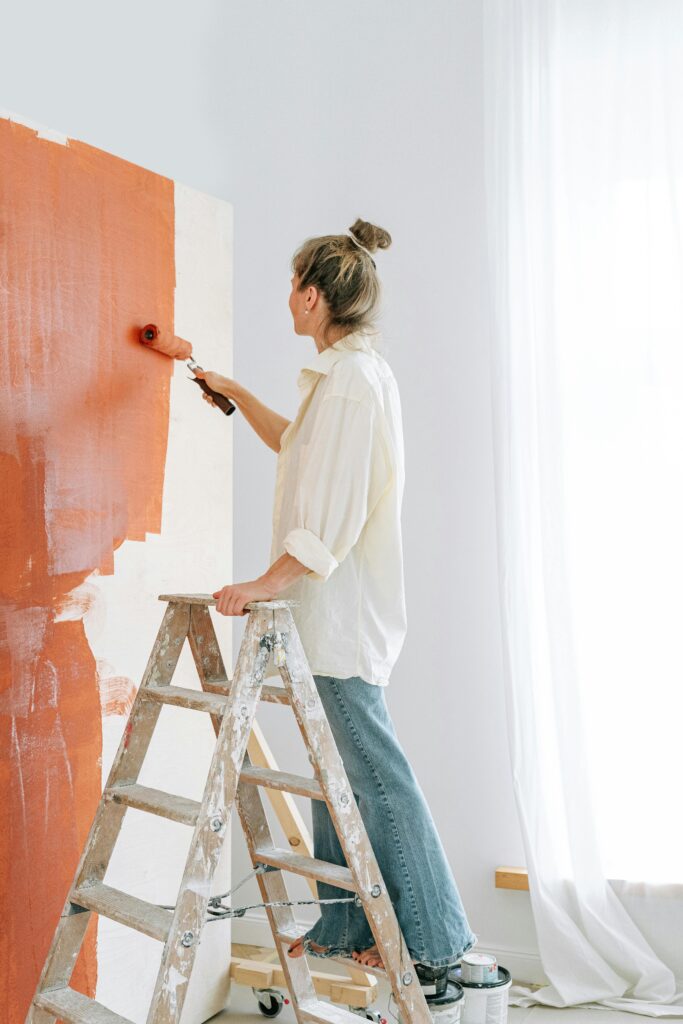 An adult woman painting a wall with red paint using a roller indoors, standing on a ladder.
