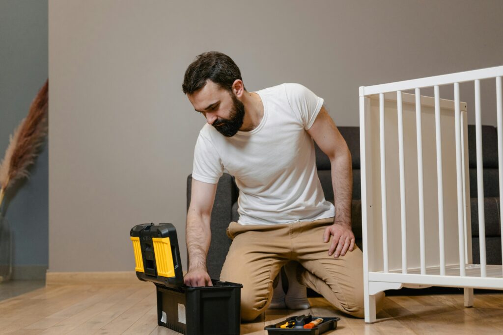 A man kneels indoors assembling a baby crib using a toolbox, wearing a white shirt and beige pants.