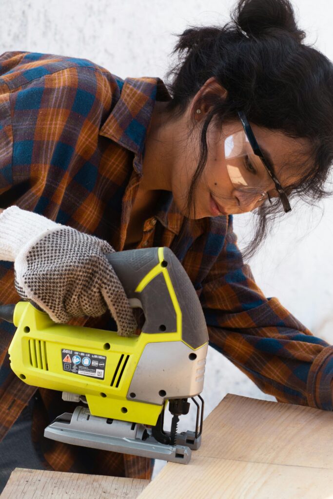 Focused woman using a jigsaw to cut wood, wearing safety goggles and gloves.